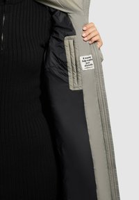 Tikkitakki harmaana mustalla vuorella, jossa on vetoketjukiinnitys ja sisäpuolella etiketti, jossa lukee "Lämmin halauksenomainen kangas."