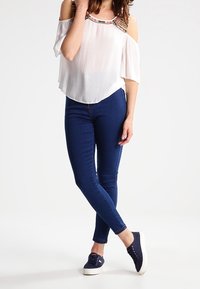 Top bianco scollato con dettagli in perline colorate, abbinato a jeans skinny blu scuro e sneakers navy. Texture liscia e design casual.