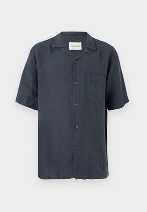 Mann steht mit Händen in den Taschen, trägt ein dunkelfarbenes kurzärmeliges Hemd mit Knöpfen, blaue Jeans und schwarze Lederschuhe auf weißem Hintergrund.