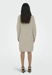 Robe en tricot à manches longues beige avec une coupe décontractée, ourlet arrondi et texture côtelée, accompagnée de sandales à talons noirs.