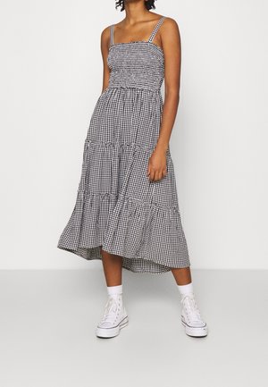 Femme portant une robe midi vichy noire et blanche à fines bretelles et des baskets montantes blanches, debout devant un fond uni.