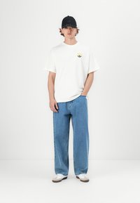 Balta kokvilnas T-kreklu ar mazu krāsainu logotipu uz krūškurvja; zila plaša denimu bikses; melna cepure; baltas un brūnas sneakers.
