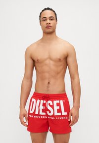 Červené plávajúce šortky vyrobené z ľahkého materiálu, s veľkým bielym logom "DIESEL" a viazaním v páse. Hladká textúra, dĺžka po strednú časť stehna.