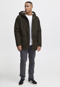 Olivgrön parkas med huva, innerfleecefoder, framdragkedja, justerbar dragsko i midjan och sidofickor; tillsammans med grå byxor och svarta skor.
