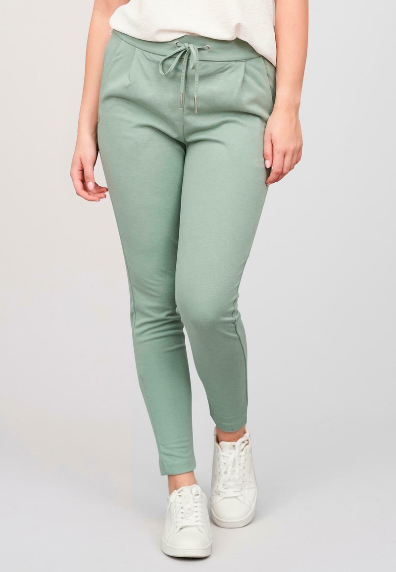 Jogging en vert clair en tissu doux ; dotés d'une taille élastique avec cordon de serrage, de poches latérales et de jambes fuselées. Portés avec des baskets blanches.