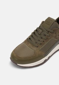Olivgrüne Sneakers aus einer Kombination von glattem Leder und strukturiertem Mesh. Akzentuiert mit flachen Schnürsenkeln und einer verstärkten Zehenkappe für Langlebigkeit.