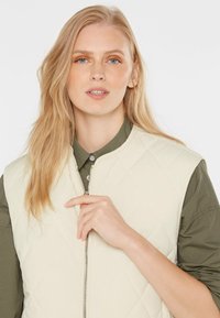 Eine blonde Frau trägt ein olivgrünes Shirt und eine cremefarbene, gesteppten Weste. Sie hält den Reißverschluss der Weste mit ihrer rechten Hand.