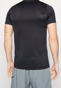 Camiseta deportiva negra con una textura suave, mangas cortas y cuello redondo. Pantalones cortos grises adicionales visibles con detalle de logo en el lateral.