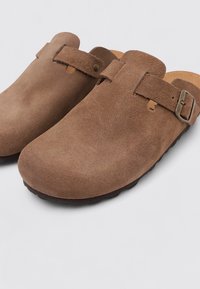 Zuecos de ante marrón con punta redondeada, que cuentan con una correa con una hebilla metálica y una superficie suave y texturizada. Suela de goma para mayor agarre.