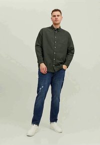Homem de pé com a mão no bolso, vestindo uma camisa de botão verde escura, jeans azuis desgastados e ténis brancos contra um fundo simples.