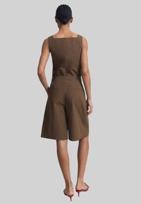 Brown, sleeveless jumpsuit with a square neckline and wide shorts. Features buttons at the waist and a smooth, textured finish.