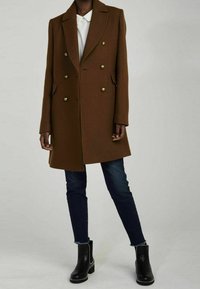 Cappotto doppiopetto marrone con bottoni dorati, rever a lancia e tasche laterali, abbinato a jeans scuri e stivaletti neri alla caviglia.