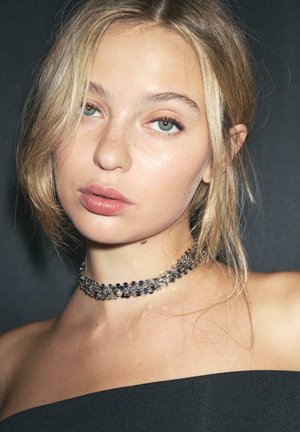 Mladá žena so svetlými blond vlasmi, svetlými pehami a zelenými očami, ktorá má na sebe trblietavý strieborný náhrdelník a čierny top bez ramienok na tmavom pozadí.