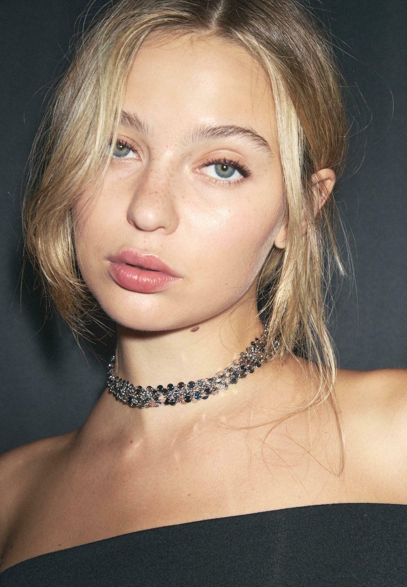 Mladá žena so svetlými blond vlasmi, svetlými pehami a zelenými očami, ktorá má na sebe trblietavý strieborný náhrdelník a čierny top bez ramienok na tmavom pozadí.