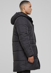 Crna puf jakna s kapuljačom, s nepropusnom teksturom, dugim krojem i zatvaračem sprijeda. Bočne džepove su jasno vidljive.