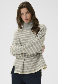 Model with long brown hair, wearing a gray and cream striped turtleneck sweater, poses with arms crossed against a plain background.