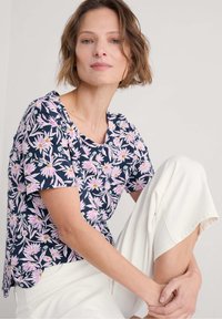 Marineblaues Kurzarmhemd mit rosa und weißen Lotusmuster, runder Ausschnitt und lockere Passform, kombiniert mit cremefarbenen Hosen.