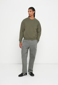 Felpa verde oliva con scollo rotondo abbinata a pantaloni cargo grigi chiari con tasche laterali e polsini alle caviglie, completata da sneakers nere.