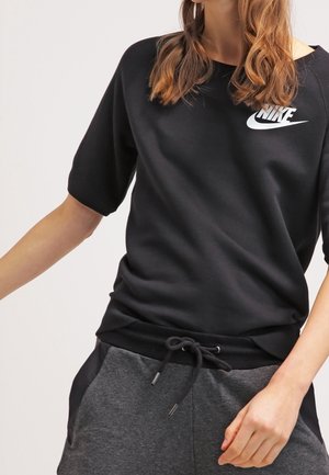 Vrouw draagt een zwart Nike-sweatshirt met korte mouwen en donkergrijze broek met trekkoord, met haar arm licht omhoog geheven.