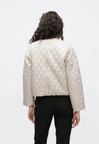 Beżowa pikowana kurtka o krótkim kroju, z przeszyciami w kształcie diamentu i szerokimi rękawami, zestawiona z czarnymi spodniami.