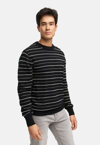 Sort sweater med vandrette hvide striber, ribbestrikket hals og manchetter, strikket tekstur. Modellen har lysegrå jeans på og står imod en ensfarvet baggrund.
