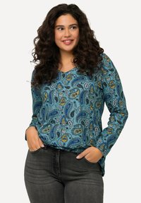 Türkises, langärmliges Oberteil mit einem Paisleymuster in Blau, Gelb und Schwarz. V-Ausschnitt-Design; getragen mit dunkelgrauen Jeans. Weicher Stoff.
