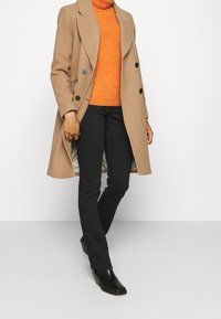 Abrigo de lana marrón con dos grandes botones negros, superpuesto a un suéter de cuello alto naranja. Pantalones negros y botas de tobillo brillantes negras completan el look.