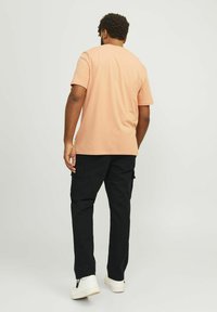 Korte mouw perzikkleurig t-shirt voor heren gecombineerd met zwarte cargobroeken. Kenmerkt zich door een relaxte pasvorm en een soepele stoftextuur. Witte sneakers zijn zichtbaar.