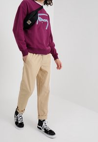 Fialová mikina s modrým potiskem, béžové kalhoty s ohrnutými manžetami a černé vysoké tenisky s bílými tkaničkami. Černá kabelka přes rameno.