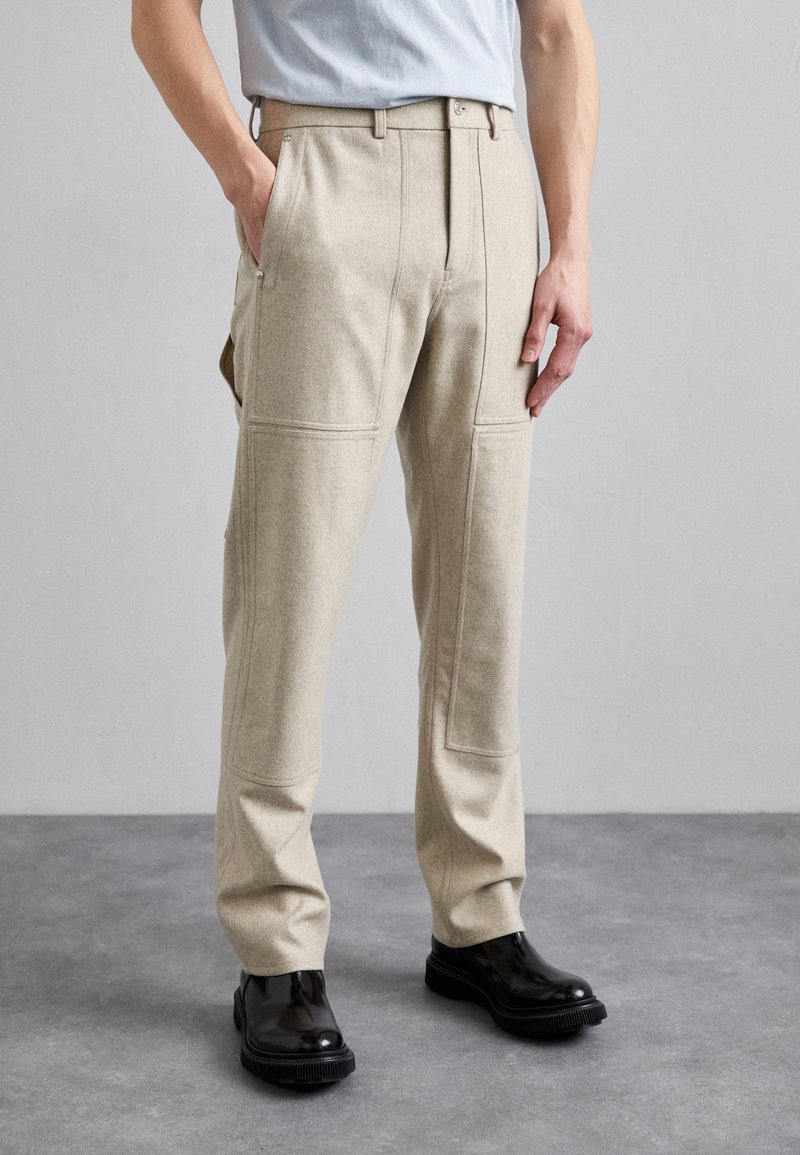 Beige wool blend trousers with flat front, side pockets, and decorative stitching; paired with glossy black shoes.