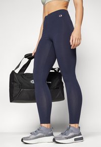 Leggings blu navy con vita alta e un leggero brillantezza, abbinati a scarpe da ginnastica grigie con design testurizzato e una borsa da palestra nera.