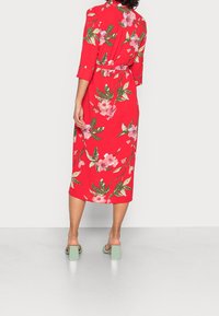 Robe rouge à fleurs avec manches trois-quarts, ceinture à la taille et longueur au genou. Elle présente des fleurs roses et des feuilles vertes, portée avec des talons blocs menthe.