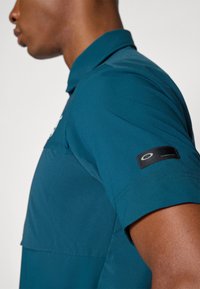 Teal short-sleeve polo shirt with a structured collar, featuring a logo patch on the sleeve and a smooth, lightweight fabric texture.