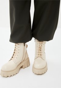 Botas de tobillo de cuero color crema con una suela gruesa y estriada, que cuentan con un cordón prominente, cremallera lateral y una textura suave.