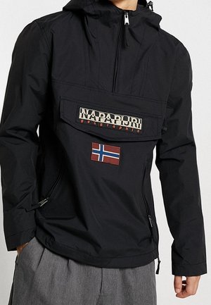 Person trägt eine schwarze Napapijri-Jacke mit Frontklappentasche und norwegischem Flaggen-Aufnäher, kombiniert mit grauer Hose.