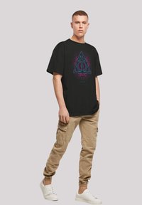 Svart T-skjorte med korte ermer som har et fargerikt trekantdesign og sirkelsymbol. Modellen har på seg kaki cargo-bukser og hvite joggesko.