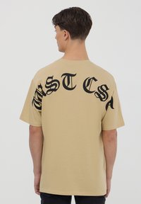 T-shirt in cotone beige con grandi lettere nere stilizzate che formano la parola "WESTCA" sul retro. Maniche corte e design collo rotondo.