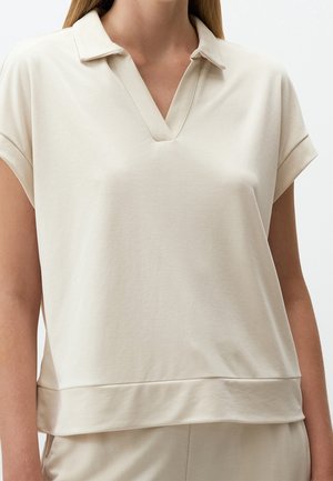 Femme portant un polo beige à manches courtes avec un col en V et un large ourlet, debout devant un fond uni.