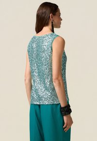 Teal paljettopp med ärmlös design, mjuk textur och rundad halsringning. Matchas med tealbyxor och svart geometrisk armband.