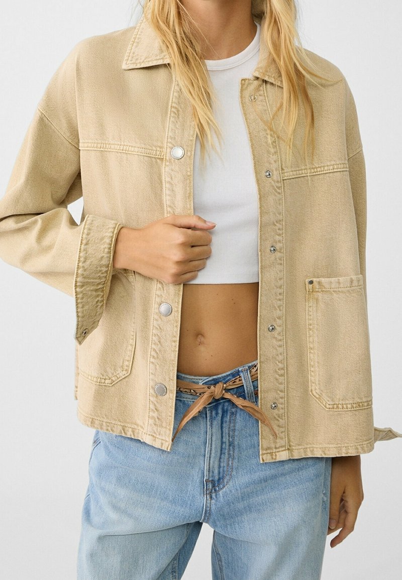 Veste en denim beige avec des manches longues, boutons en argent, poche poitrine et coupe ample, portée par-dessus un top blanc et un jean bleu clair.