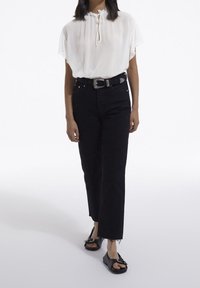 Vit, kortärmad blus med en halsknut, matchad med hög midja svarta raka jeans och svarta slip-on sandaler med en rosettdetalj.