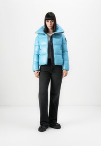 Vrouw staat rechtop met het gezicht naar voren, gekleed in een lichtblauwe pufferjack, zwarte broek, zwarte platform schoenen en een witte shirt tegen een witte achtergrond.