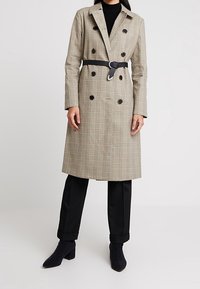 Double-breasted trench coat in beige plaid fabric with a belted waist, notched lapels, and black buttons. Paired with black trousers and ankle boots.
