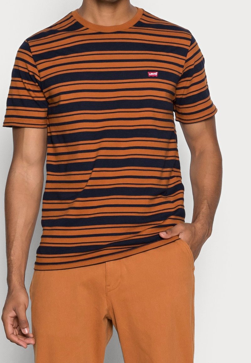Homme portant un t-shirt à manches courtes rayé orange et noir et un pantalon orange uni, avec une main dans la poche et l'autre détendue.