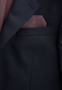 Poche d'une veste de costume bleu marine avec un mouchoir de poche bordeaux à motifs plié à l'intérieur.