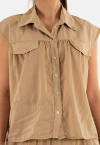 Blusa beige de manga corta con cierre de botones en la parte delantera, dos bolsillos en el pecho y un diseño con pliegues, hecha de tela ligera. Cuenta con botones plateados.