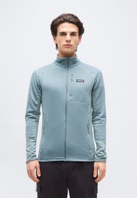 Νεαρός άνδρας με σκούρα μαλλιά φοράει ανοιχτό μπλε τζάκετ με φερμουάρ και μαύρο παντελόνι, στέκεται μπροστά σε απλό λευκό φόντο.