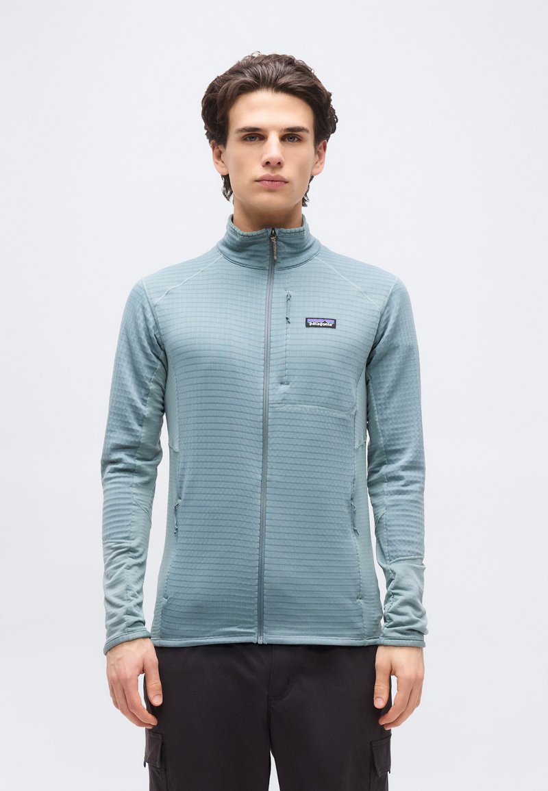 Νεαρός άνδρας με σκούρα μαλλιά φοράει ανοιχτό μπλε τζάκετ με φερμουάρ και μαύρο παντελόνι, στέκεται μπροστά σε απλό λευκό φόντο.