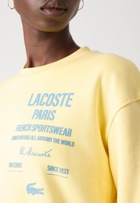 Gros plan d'une personne portant un sweat-shirt jaune avec le texte et le logo bleu Lacoste Paris French sportswear à l'avant.