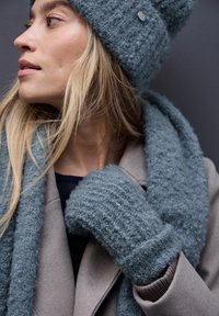 Un gorro de punto azul claro con textura, con un botón, que combina con una bufanda y unos guantes, todo ello sobre un abrigo gris y una blusa azul marino.